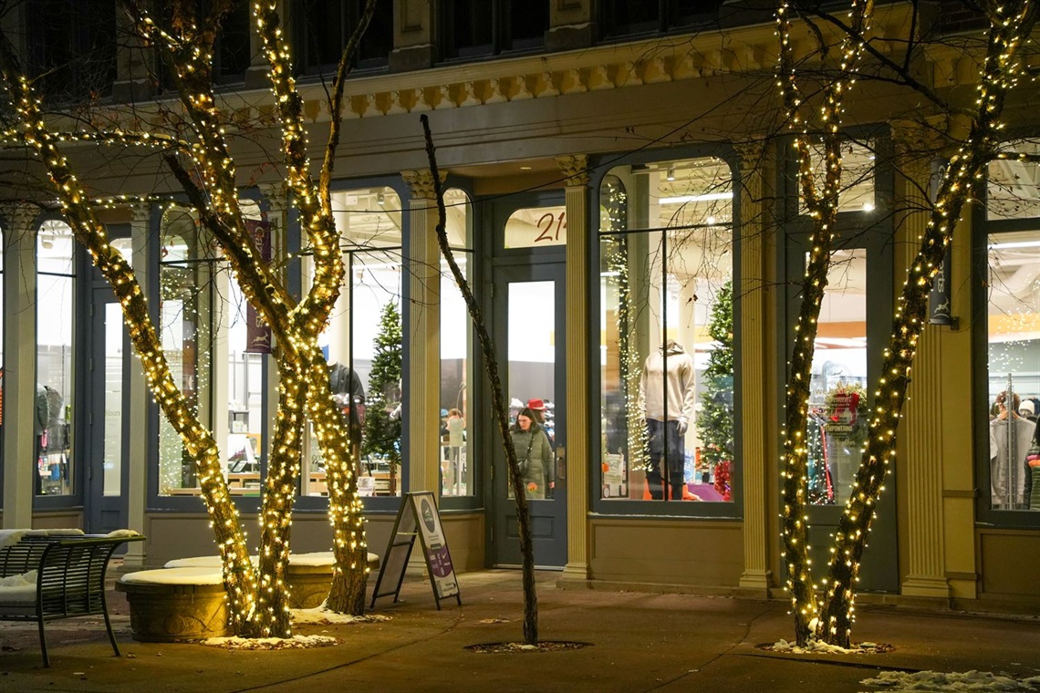 A photo of a downtown shop at night with holiday lights on the trees in front of it and people shopping inside. 