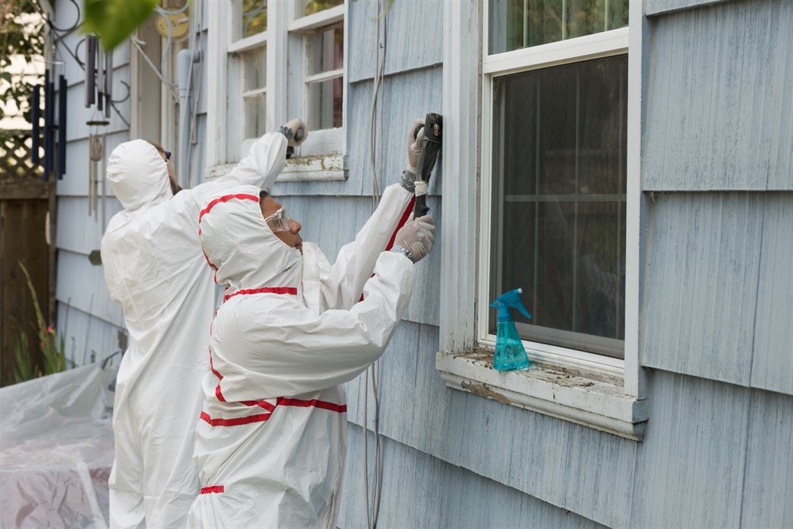 photo of two lead abatement workers in white protective gear performing lead abatement on a blue home. 