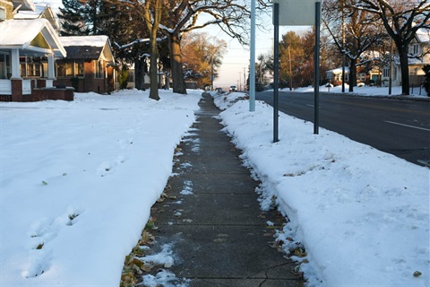 A photo of a neighborhood sidewalk with snow on the ground and the sidewalk cleared of snow. 